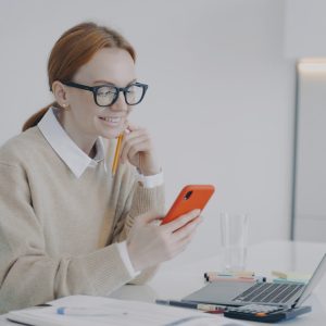 Smiling young woman is texting at workplace or at class. Girl is being distracted at work in office.