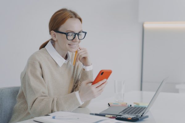 Smiling young woman is texting at workplace or at class. Girl is being distracted at work in office.